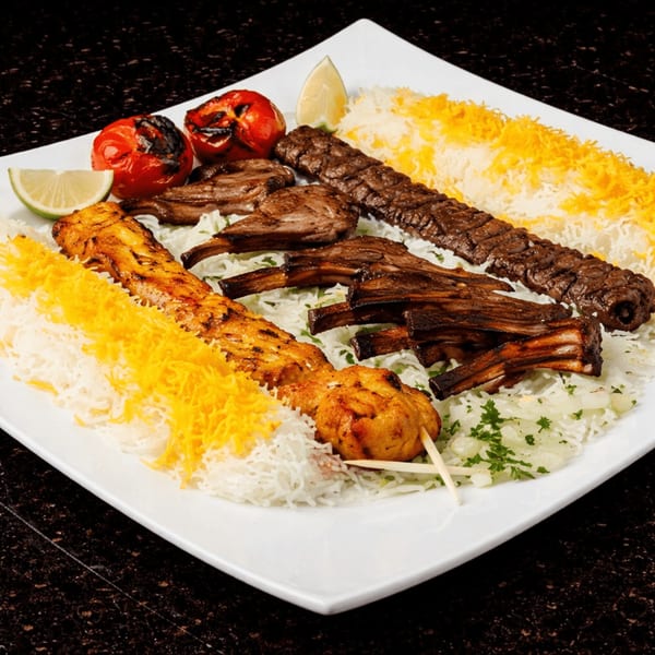 A white plate of Persian kabobs, including koobideh, lamb chops, and chicken, with rice and grilled tomatoes.